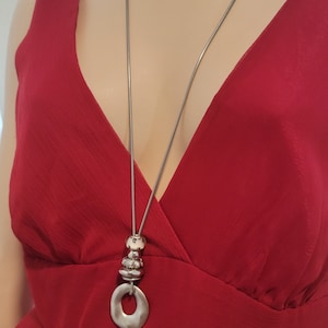 May include: A silver chain necklace with a large, round pendant and three smaller round beads. The pendant has a hole in the center. The necklace is on a mannequin wearing a red dress.