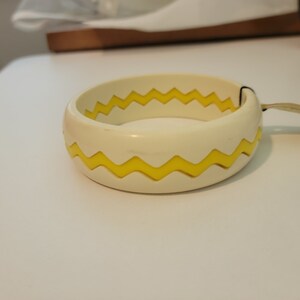 Könnte beinhalten: Cremefarbenes Armreif-Armband mit gelbem Zickzackmuster. Das Armband besteht aus einem festen Material und hat eine glatte Oberfläche. Ein kleines schwarzes Etikett und ein beigefarbener Riemen sind am Armband befestigt.