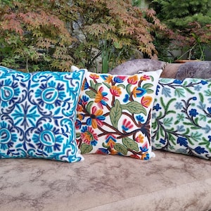 May include: Three decorative throw pillows with embroidered floral designs. One pillow features a blue and white pattern, another has colorful flowers, and the third has green and blue leaves. The pillows are on a brown couch.