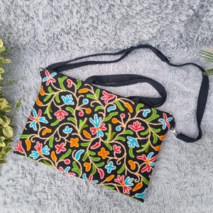 Könnte beinhalten: Schwarze Umhängetasche mit farbenfrohem Blumenstickerei-Design. Die Tasche hat einen schwarzen, verstellbaren Riemen und einen Reißverschluss. Die Stickerei umfasst rosa, blaue, orange und grüne Blumen und Blätter.