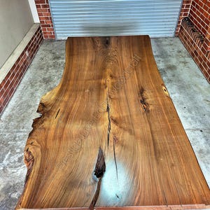 May include: A large, live edge wooden table top with a natural wood finish. The table top has a unique, irregular shape and a light blue resin fill in a crack.