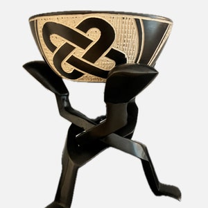 Op de afbeelding: Een zwarte keramische kom met een wit geometrisch ontwerp staat op een zwarte metalen standaard in de vorm van een persoon die de kom draagt.