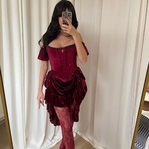 Puede incluir: Un vestido cors&eacute; de terciopelo burdeos con mangas ca&iacute;das y una falda con volantes. El vestido presenta un corpi&ntilde;o ajustado y se combina con medias de encaje y tacones a juego. El atuendo es un ejemplo llamativo de moda g&oacute;tica o de inspiraci&oacute;n vintage.