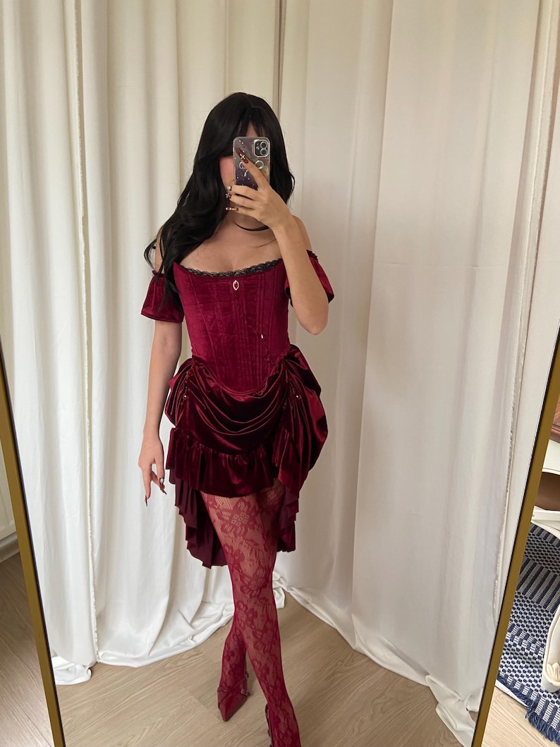 Puede incluir: Un vestido cors&eacute; de terciopelo burdeos con mangas ca&iacute;das y una falda con volantes. El vestido presenta un corpi&ntilde;o ajustado y se combina con medias de encaje y tacones a juego. El atuendo es un ejemplo llamativo de moda g&oacute;tica o de inspiraci&oacute;n vintage.