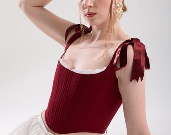 May include: A burgundy corset top with ribbon shoulder ties and a white lace trim. The top is paired with a white skirt. The model is wearing gold earrings and a lace glove.