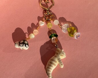 May include: A gold heart-shaped keychain with a variety of charms, including a turtle, a cat, and flowers. The charms are made of ceramic and glass and are strung on a gold chain.