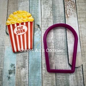 May include: A red and white striped cookie cutter shaped like a popcorn bucket with the words "Pop Corn" on the front. A purple cookie cutter shaped like a half circle is also in the image.