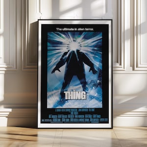 May include: A framed movie poster for "The Thing" features a dark figure with a bright light emanating from its head. The title "THING" is prominently displayed in white, with the tagline "The ultimate in alien terror." The poster is in a black frame.