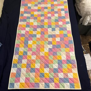 Pode incluir: Uma colcha de retalhos colorida com um padrão repetitivo de quadrados em tons pastel de rosa, amarelo, azul, verde e lavanda. Alguns quadrados apresentam uma estampa floral. A colcha tem uma borda branca e está sobre uma superfície azul marinho.
