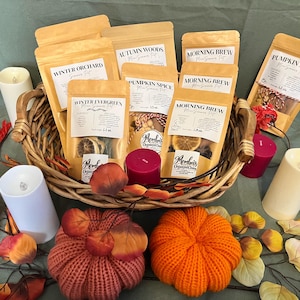 May include: A woven wicker basket filled with several tan pouches labeled Winter Orchard, Autumn Woods, Morning Brew, and Pumpkin Spice. Each pouch features a clear window displaying dried fruit and spices, accompanied by candles and fabric pumpkins.