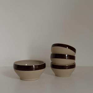 Puede incluir: Un juego de cuatro cuencos de cerámica beige con una banda marrón oscuro alrededor del borde y un cuenco solo a la izquierda. Los cuencos están apilados sobre una superficie blanca. Cada cuenco mide unos 12 cm de diámetro.