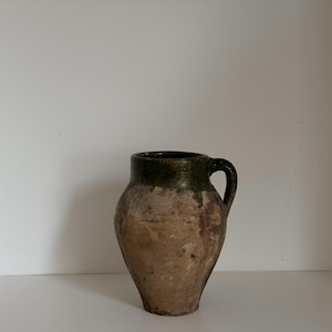 May include: A rustic, antique-style pottery jug with a handle. The jug has a bulbous body and a dark green rim. The exterior has a weathered, earthy brown color, suggesting age and use. The jug is likely used for decorative purposes.