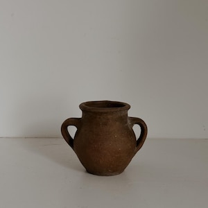May include: A small, brown, two-handled ceramic vase. The vase has a rounded body and a slightly flared rim. The handles are loop-shaped and attached to the sides of the vase. The surface has a textured appearance.