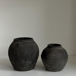 Pode incluir: Dois vasos redondos de cerâmica cinza escuro de tamanhos diferentes. O vaso maior está à esquerda e o menor à direita. Ambos os vasos têm uma parte superior larga e aberta e uma superfície texturizada. Os vasos são exibidos em uma superfície branca.