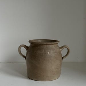 May include: A tan ceramic jar with two loop handles. The jar has a wide opening and a slightly textured surface. The overall shape is cylindrical with a subtle curve. The jar is likely for decorative or functional use.