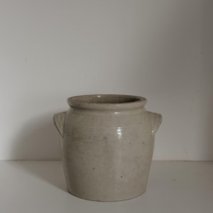 May include: A light beige stoneware crock with two small handles and a slightly flared rim. The antique jar has horizontal bands around the middle and a smooth, glossy finish. The neutral colour and classic shape make it a versatile decorative piece.