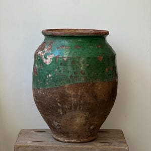 以下が含まれることがあります： 大きく丸みを帯びた形状の、アンティークの陶器製の壺。上半分は鮮やかな緑色、下半分は風化した茶色に塗装されています。素朴な木製のスツールの上に置かれ、そのサイズと質感を際立たせています。