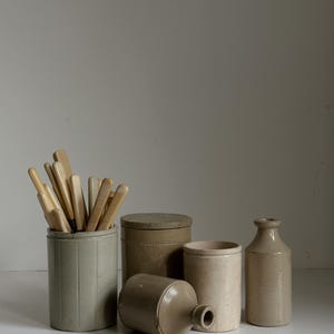 Puede incluir: Colección de cerámica antigua de gres en varias formas y tamaños. Incluye un tarro cilíndrico con utensilios de madera, un recipiente con tapa, un cilindro pequeño abierto y una botella. Las piezas son de color beige y gris.