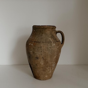 May include: An antique earthenware jug with a single handle. The jug has a bulbous body and a narrow neck. The exterior is a mottled brown with visible texture and a horizontal line near the top. The jug is likely used for storing liquids.
