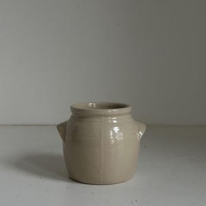 Puede incluir: Un pequeño jarrón de cerámica beige con dos pequeñas asas a los lados. El jarrón tiene un cuerpo redondeado y un borde ligeramente ensanchado. La superficie tiene un acabado mate y parece hecho a mano. El jarrón está sobre una superficie blanca.