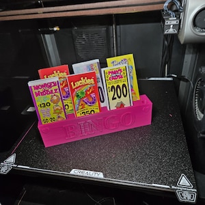 Puede incluir: Un soporte rosa impreso en 3D para tarjetas de bingo con la palabra "BINGO" en la parte delantera. El soporte contiene varios boletos de lotería para raspar de colores. Los boletos tienen varios diseños y texto, incluyendo "Luckies" y premios.