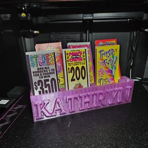 May include: A purple 3D-printed organizer with the name "KATHRYN" on the front, holding several scratch-off lottery tickets. The tickets have colorful designs and text, including "STORES," "PAWS for the Cause," and "Triple Twist."