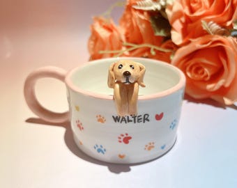 May include: A white ceramic mug with a light pink handle and rim, decorated with colorful paw prints and the name "WALTER". A small, tan-colored dog figurine is perched on the rim. The background features orange roses.