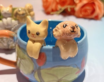 Può includere: Una tazza di ceramica blu con una statuina di gatto e cane attaccata al bordo. La tazza presenta un motivo floreale con accenti arancioni e verdi. Il gatto e il cane sono giallo chiaro e marrone chiaro.