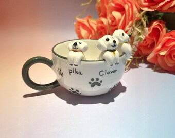May include: White ceramic teacup with three small white dog figurines perched on the rim. The cup has a green handle and the names "Pika" and "Clover" are written on the side, along with paw prints. Orange roses are in the background.