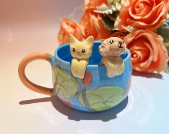 Può includere: Una tazza in ceramica con interno blu e manico arancione chiaro. La tazza presenta un gatto giallo e una statuina di cane marrone chiaro sul bordo. L'esterno è decorato con motivi floreali verdi e blu. Rose arancioni sullo sfondo.