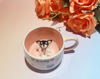 Può includere: Una tazza in ceramica bianca con interno e manico rosa, con una piccola statuina di chihuahua con ali d'angelo all'interno. La tazza è decorata con cuori e la parola "Birdy". Rose arancioni sullo sfondo.