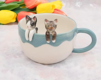 Può includere: Una tazza in ceramica con manico e bordo azzurro, con due piccole figure di gatto appollaiate sul bordo. La tazza è bianca con un motivo a onde blu. Tulipani gialli e rosa sullo sfondo.