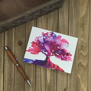 May include: A greeting card featuring a watercolor tree design in shades of pink, purple, and blue. A wooden pen with silver accents rests beside the card on a wooden surface. A decorative wooden box is in the background.