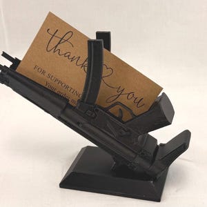 May include: Black gun-shaped business card holder with a brown card that says "Thank you" in cursive. The holder is angled upwards, with a rectangular base and a slot to hold the card. The card also says "FOR SUPPORTING Your order".