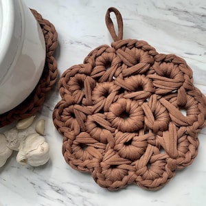 Op de afbeelding: Een bruine gehaakte pot onderzetter met een cirkelvormig ontwerp. De pot onderzetter is gemaakt van een zachte, geweven stof en heeft een uniek, ingewikkeld patroon.
