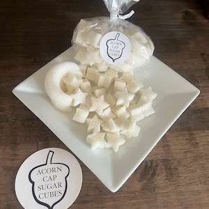 May include: A white square plate holds star-shaped and cube-shaped sugar cubes, along with a sugar ring. A clear bag of sugar cubes is tied with a ribbon. A round sticker with the text "Acorn Cap Sugar Cubes" is visible.