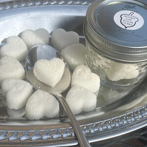 May include: A silver tray with a glass jar of white sugar cubes. The jar has a label that reads "Acorn Cap Sugar Cubes". The tray also has a silver spoon and several white heart-shaped sugar cubes.