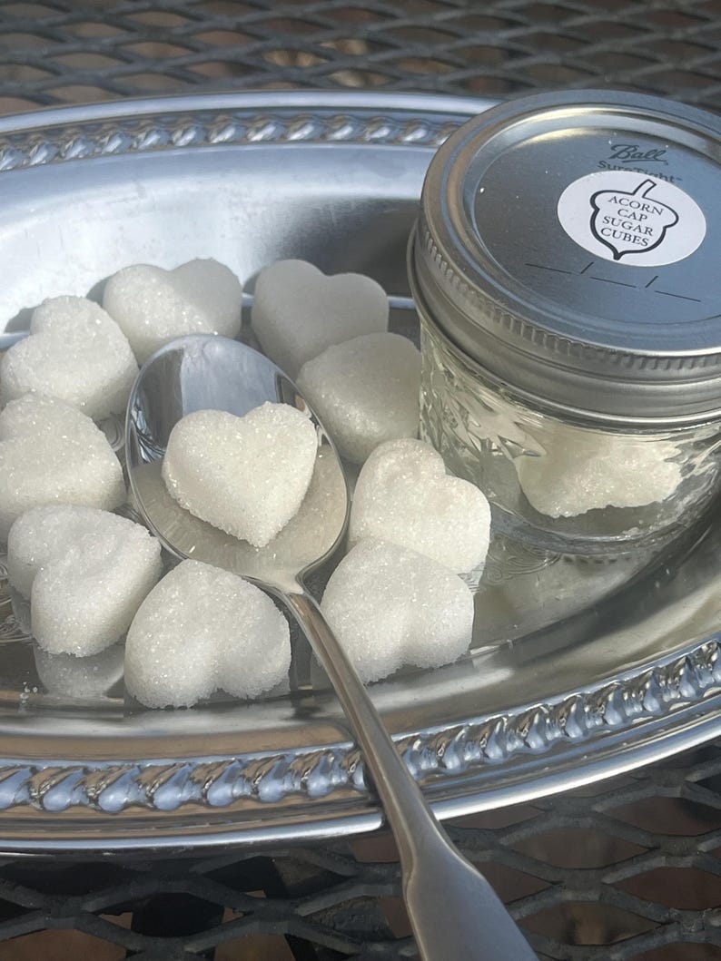 May include: A silver tray with a glass jar of white sugar cubes. The jar has a label that reads "Acorn Cap Sugar Cubes". The tray also has a silver spoon and several white heart-shaped sugar cubes.
