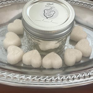 May include: A glass jar with a silver lid filled with white heart-shaped sugar cubes. The jar has a label that reads "Ball SureTight Acorn Gap Sugar Cubes". The sugar cubes are on a silver tray.
