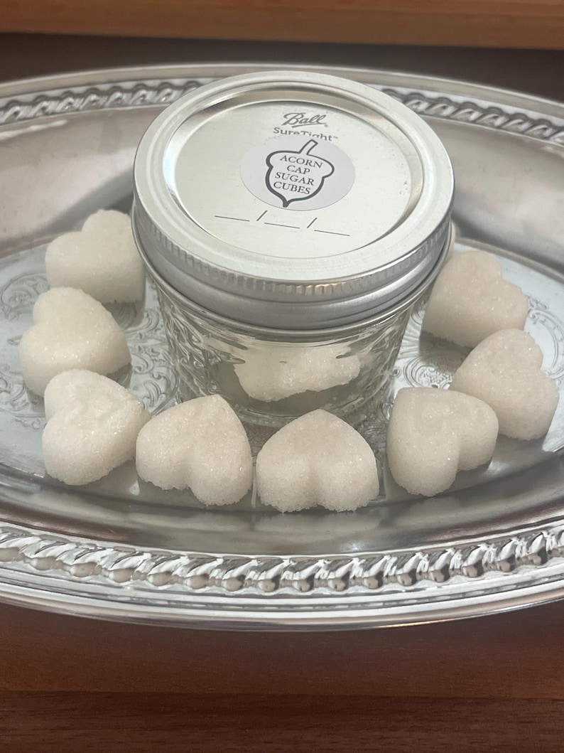 May include: A glass jar with a silver lid filled with white heart-shaped sugar cubes. The jar has a label that reads "Ball SureTight Acorn Gap Sugar Cubes". The sugar cubes are on a silver tray.