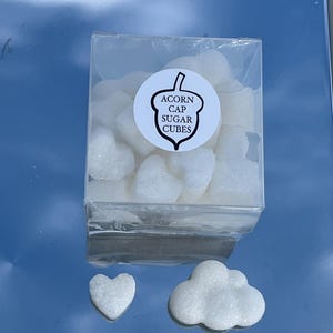 Puede incluir: Un recipiente de plástico transparente lleno de cubos de azúcar blancos con forma de corazón. El recipiente tiene una etiqueta que dice "Acorn Cap Sugar Cubes". También hay dos cubos de azúcar fuera del recipiente, uno con forma de corazón y otro con forma de nube.