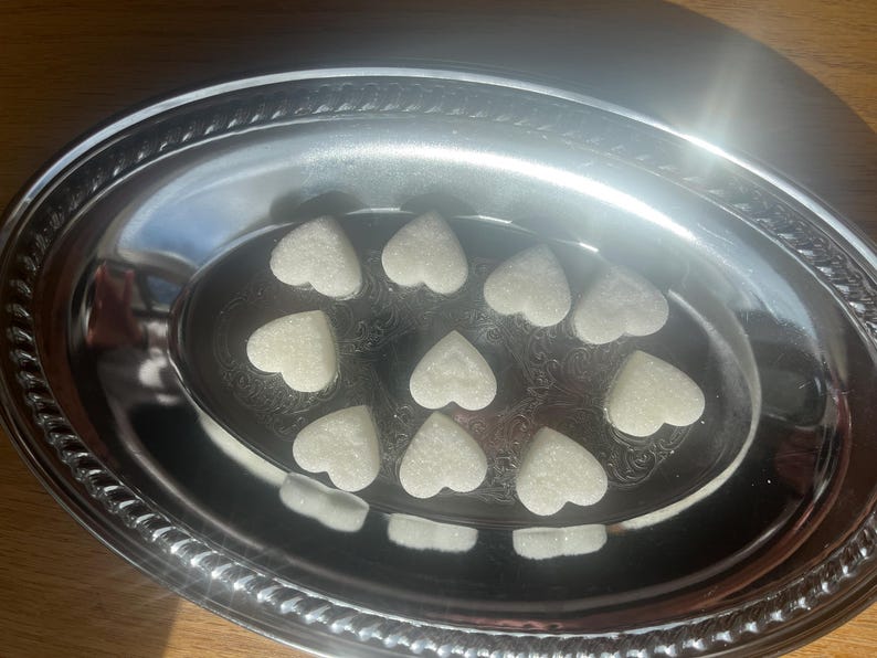 May include: A silver oval serving dish with a decorative border holds 10 white heart-shaped sugar cubes.