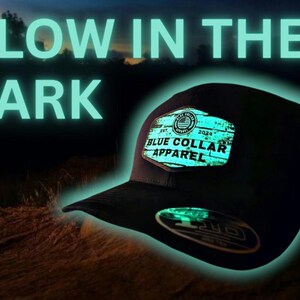 Könnte beinhalten: Eine schwarze Baseballkappe mit einem im Dunkeln leuchtenden Patch, auf dem in weißer Schrift "Blue Collar Apparel" steht. Der Patch hat eine Holzmaserung und eine rot-weiß-blaue amerikanische Flagge in der oberen linken Ecke. Der Text "Est. 2020" befindet sich in der oberen rechten Ecke des Patches.