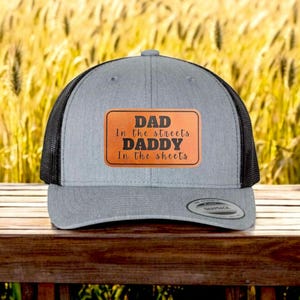 Puede incluir: Gorra de camionero gris y negro con un parche de cuero marrón que dice "Dad in the streets Daddy in the sheets".
