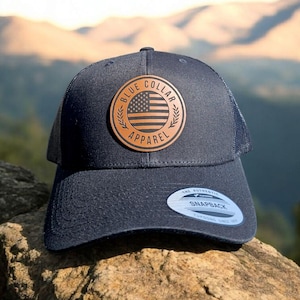Könnte beinhalten: Eine schwarze und blaue Trucker-Kappe mit einem braunen Leder-Patch, auf dem "Blue Collar Apparel" steht und eine kleine amerikanische Flagge abgebildet ist. Die Kappe liegt auf einem Stein.