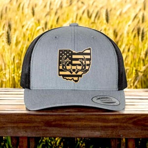 May include: A gray and black trucker hat with a patch of the state of Ohio with an American flag design and a pair of antlers. The hat is sitting on a wooden surface.