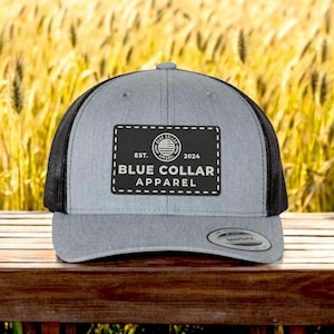 Könnte beinhalten: Eine graue und schwarze Trucker-Kappe mit einem schwarzen Patch, auf dem "EST. 2024 BLUE COLLAR APPAREL" steht, mit einem kleinen Kreislogo über dem Text.