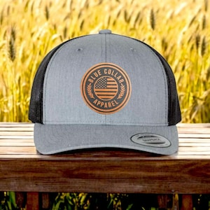Könnte beinhalten: Eine graue und schwarze Trucker-Kappe mit einem braunen Leder-Patch, auf dem "Blue Collar Apparel" steht und eine amerikanische Flagge abgebildet ist.
