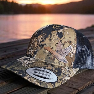 May include: Camouflage baseball cap with a brown leather patch of a flying duck. The cap has a black mesh back and a curved brim. The text "THE AUTHENTIC SNAPBACK YUPOONG SINCE 1974" is on a silver oval patch.