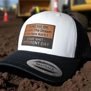 May include: A black and white trucker hat with a white front panel and a black brim and mesh back. The hat features a brown leather patch with the text "PLEASE PICK ONE" and three phrases: "Livin' The Dream", "I'm Here Ain't I", and "SAME SHIT - DIFFERENT DAY".